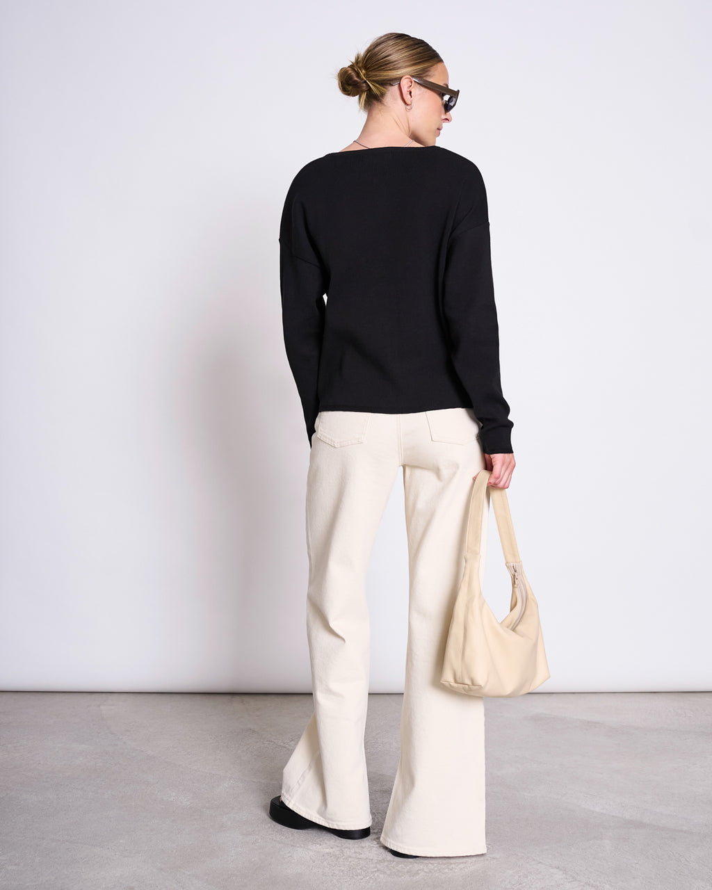 A woman stands with her back to the camera wearing the jan-n-june LIGHT KNIT JUMPER WILLIAMSBURG BLACK GOTS, cream wide-leg organic cotton knit pants, black shoes, a beige handbag, sunglasses, and her hair styled in a neat bun.