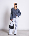 A woman stands against a plain background wearing black sunglasses, white wide-leg pants, black sandals, a black shoulder bag, and the jan-n-june CARDIGAN ANJO TURBULENCE GOTS in an oversized dark gray knit.