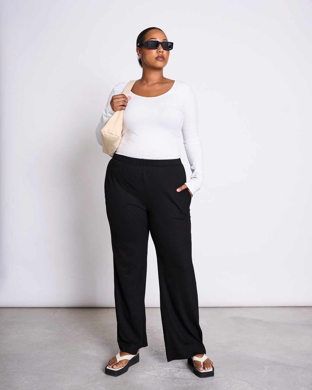 A woman in jan-n-junes BALLERINA LONGSLEEVE LEON SOFT WHITE GOTS top, black wide-leg pants, sunglasses, and sandals stands against a plain background, holding a beige shoulder bag with one hand in her pocket.