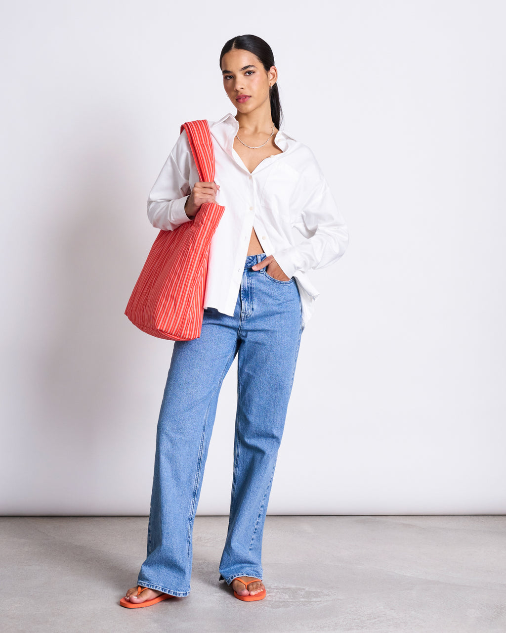 A woman with long dark hair and a confident pose wears the jan-n-june OVERSIZED SHIRT MAMRO WHITE, styled with blue jeans, orange sandals, and a large red striped shoulder bag against a plain backdrop.