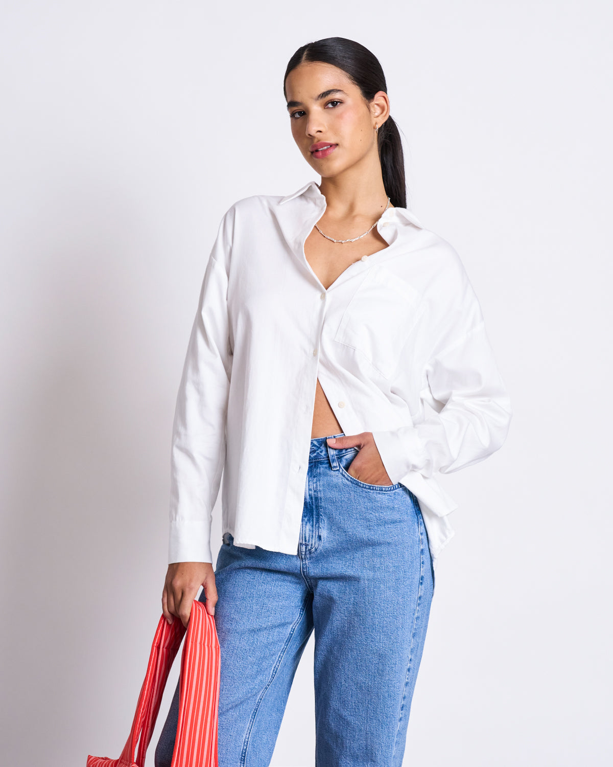 A woman models the jan-n-june OVERSIZED SHIRT MAMRO WHITE with blue jeans, standing against a white background and holding a red striped bag. Her long dark hair is pulled back as she looks confidently at the camera.