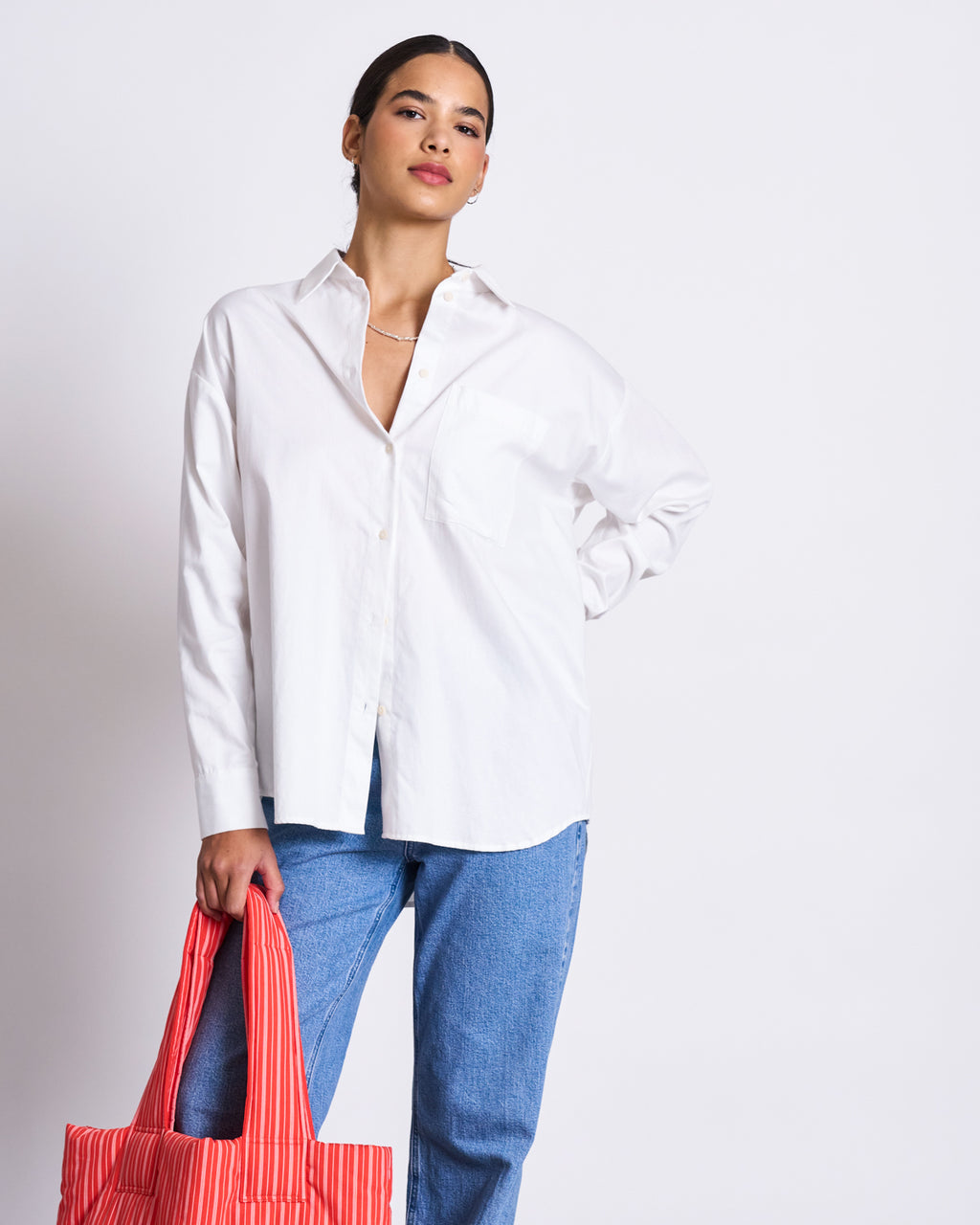 A person stands against a plain background wearing jan-n-junes OVERSIZED SHIRT MAMRO WHITE and blue jeans, holding a red and white striped tote bag in their left hand.