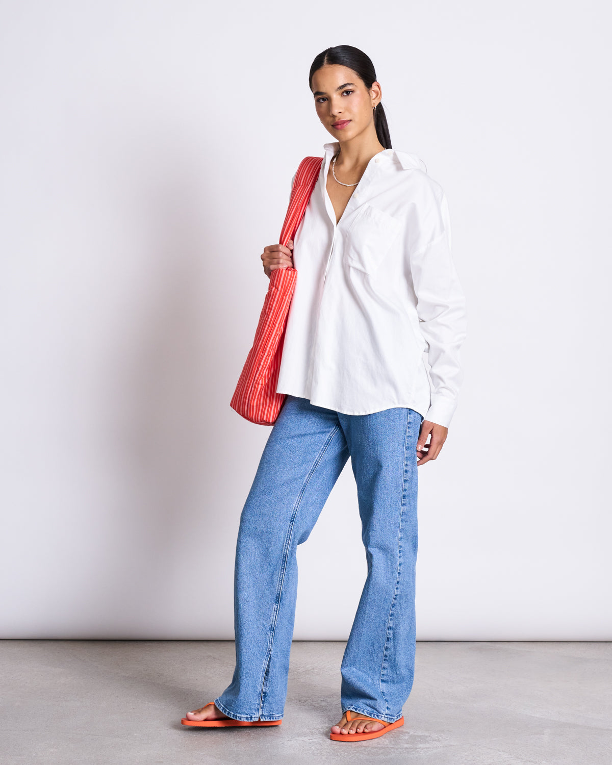 A woman stands against a plain background wearing the jan-n-june OVERSIZED SHIRT MAMRO WHITE, blue jeans, orange sandals, and carries a red-striped shoulder bag.