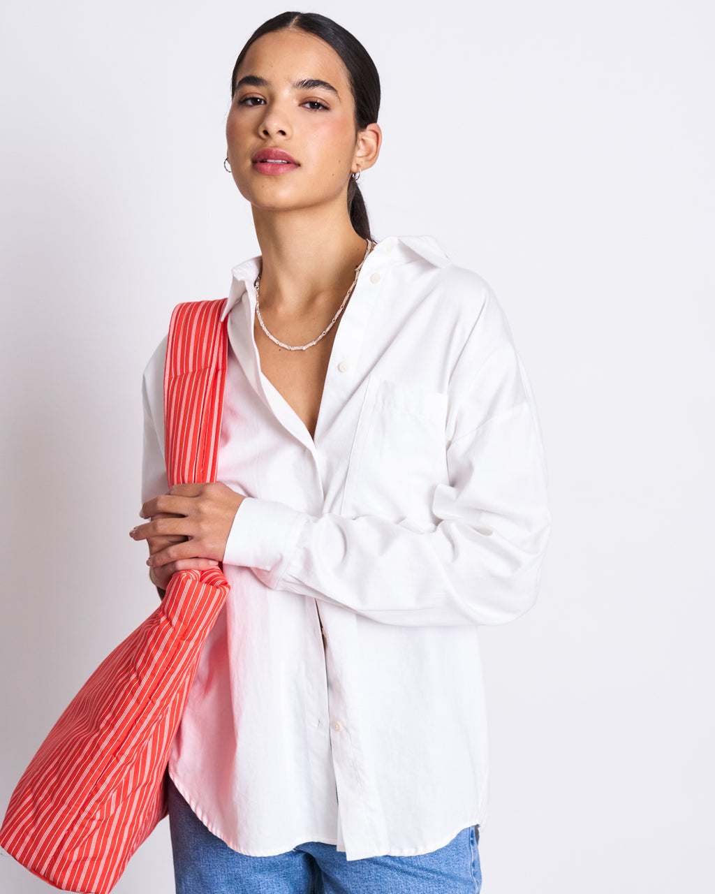 A woman with dark hair pulled back wears the jan-n-june OVERSIZED SHIRT MAMRO WHITE and blue jeans, accessorized with a simple necklace, earrings, and a red-and-white striped bag against a plain background.