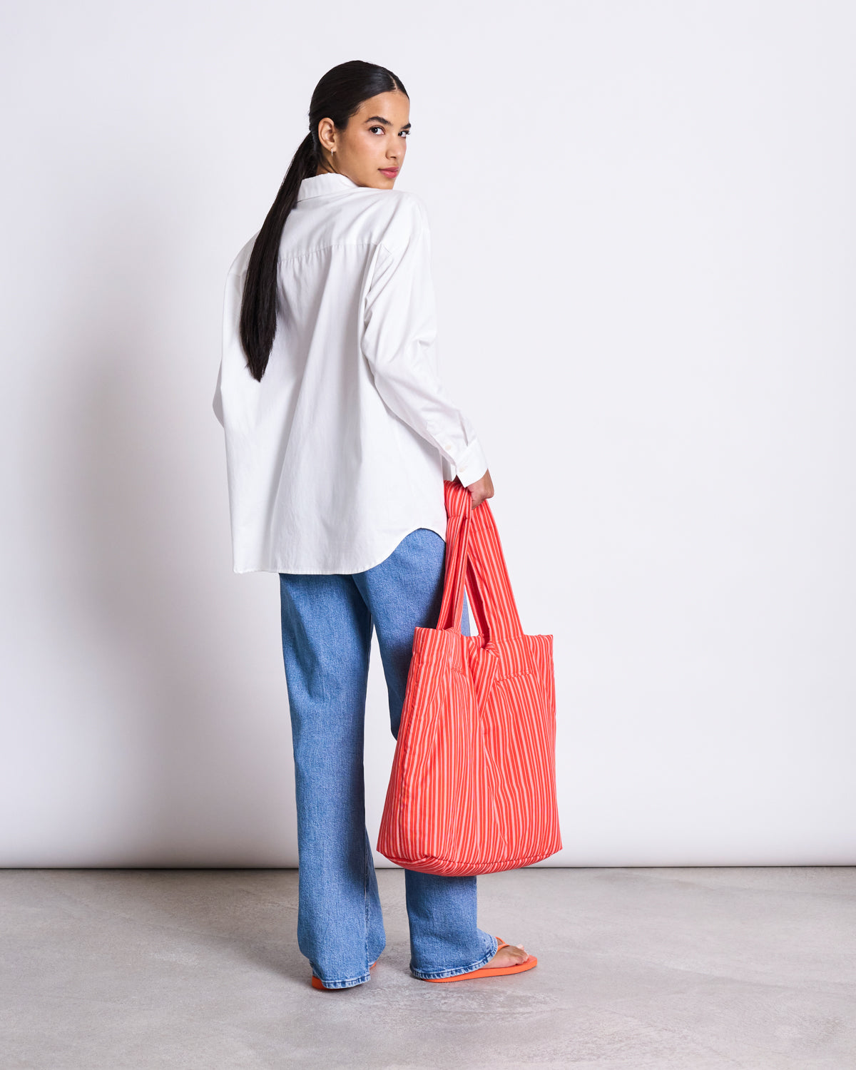 A woman faces away, turning to the camera in a jan-n-june OVERSIZED SHIRT MAMRO WHITE, blue jeans, orange sandals, and a red striped tote bag. The background is plain and light-colored.