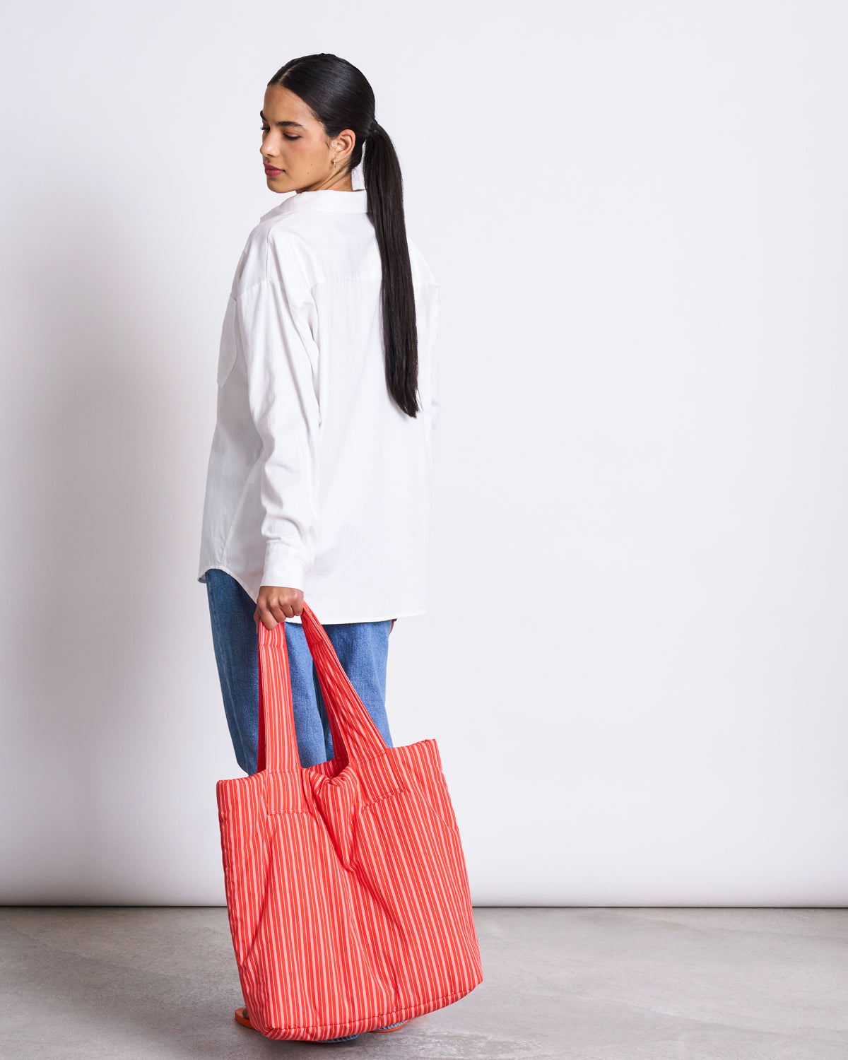 A woman with long dark hair in a ponytail, wearing a white shirt and blue jeans, stands sideways holding the jan-n-june PADDED SHOPPER SMOL CHERRY RED WHITE STRIPED. She looks over her shoulder against a plain light background.