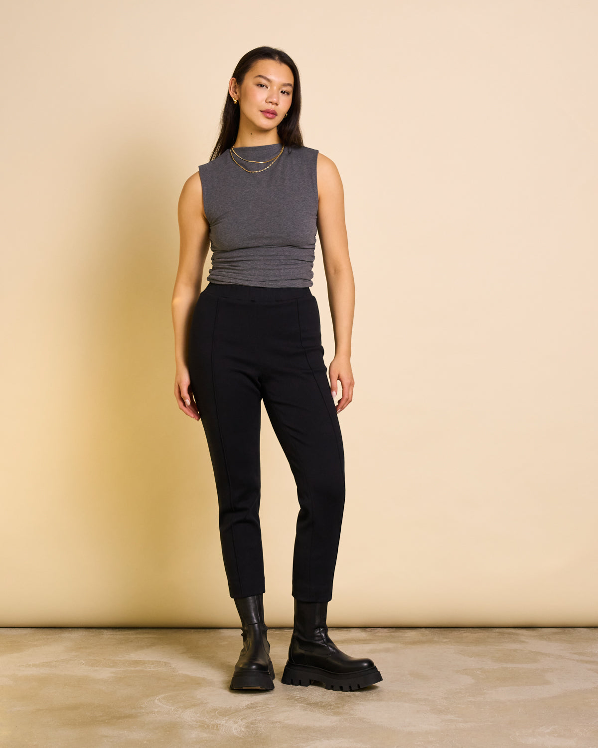 A woman with straight dark hair poses confidently against a beige background, wearing jan-n-june PANTS KAMA BLACK GOTS high-waisted organic cotton pants, a sleeveless gray top, black chunky boots, and layered gold necklaces.