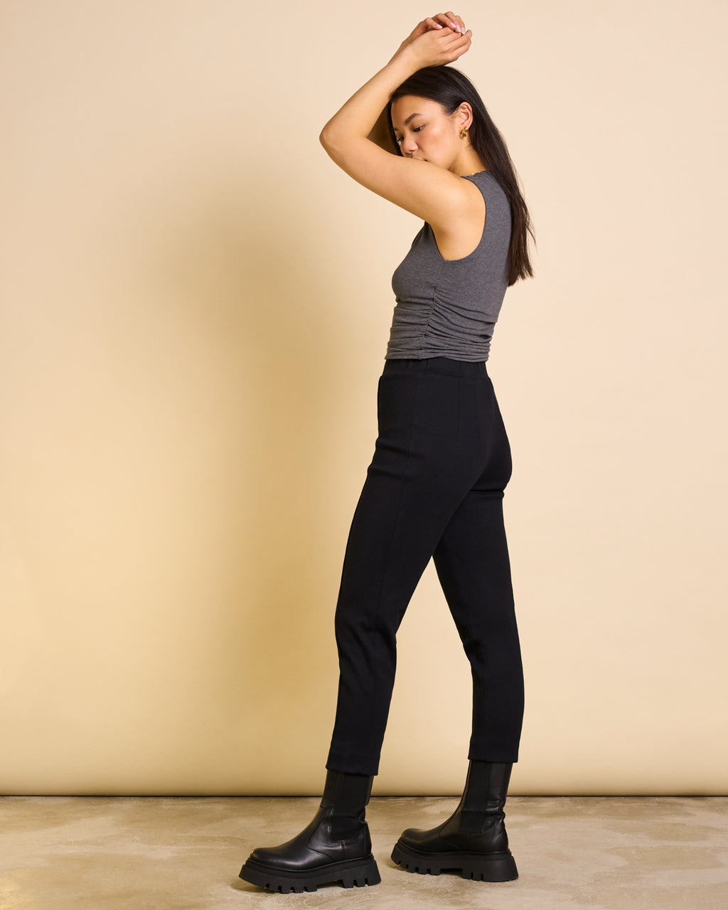 A woman stands sideways with arms raised, wearing a sleeveless gray top, jan-n-june’s PANTS KAMA BLACK GOTS, and black chunky ankle boots against a beige background.
