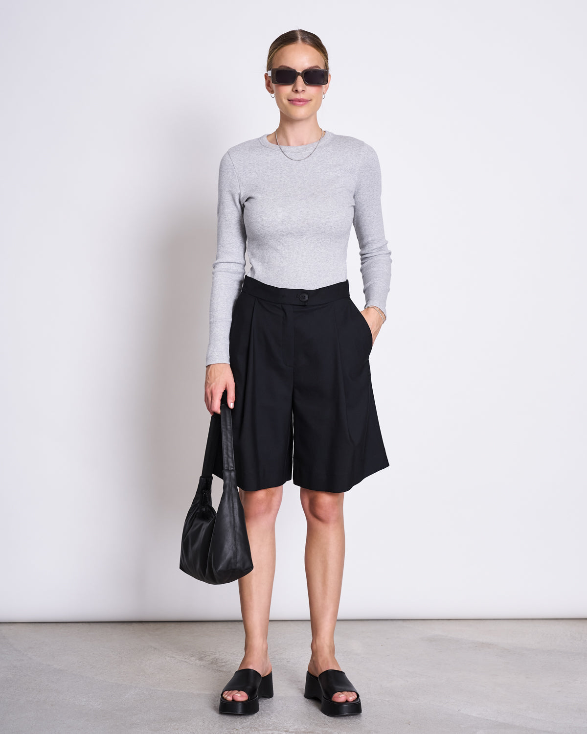 A woman wears the jan-n-june RIB LONGSLEEVE MURI LIGHT GREY GOTS with sunglasses, black knee-length shorts, black platform sandals, and a black handbag. She poses with one hand in her pocket—perfect for chic layering.