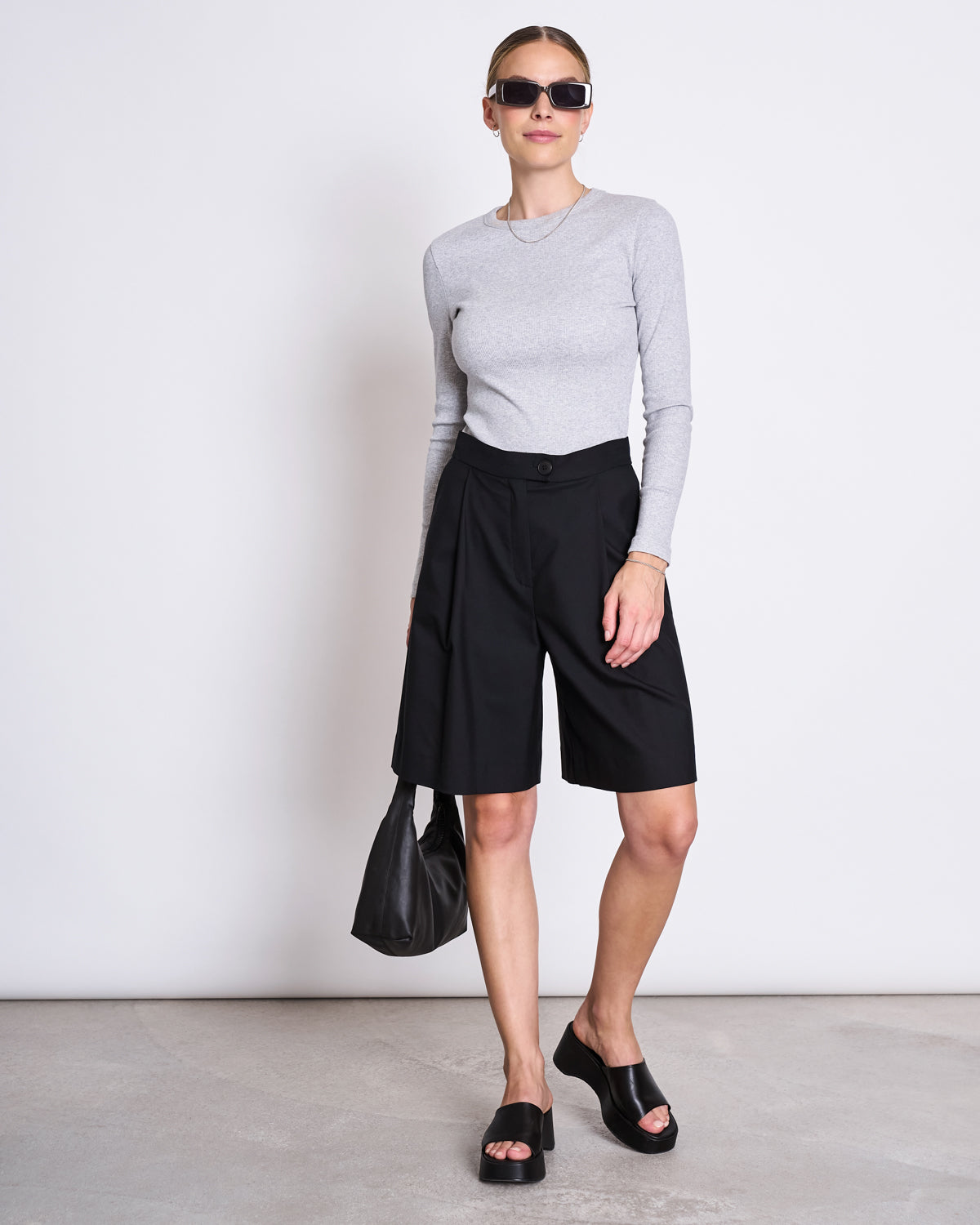 A woman in sunglasses and the jan-n-june RIB LONGSLEEVE MURI LIGHT GREY GOTS top, styled with black knee-length shorts, black platform sandals, and a black handbag, stands against a plain white wall.