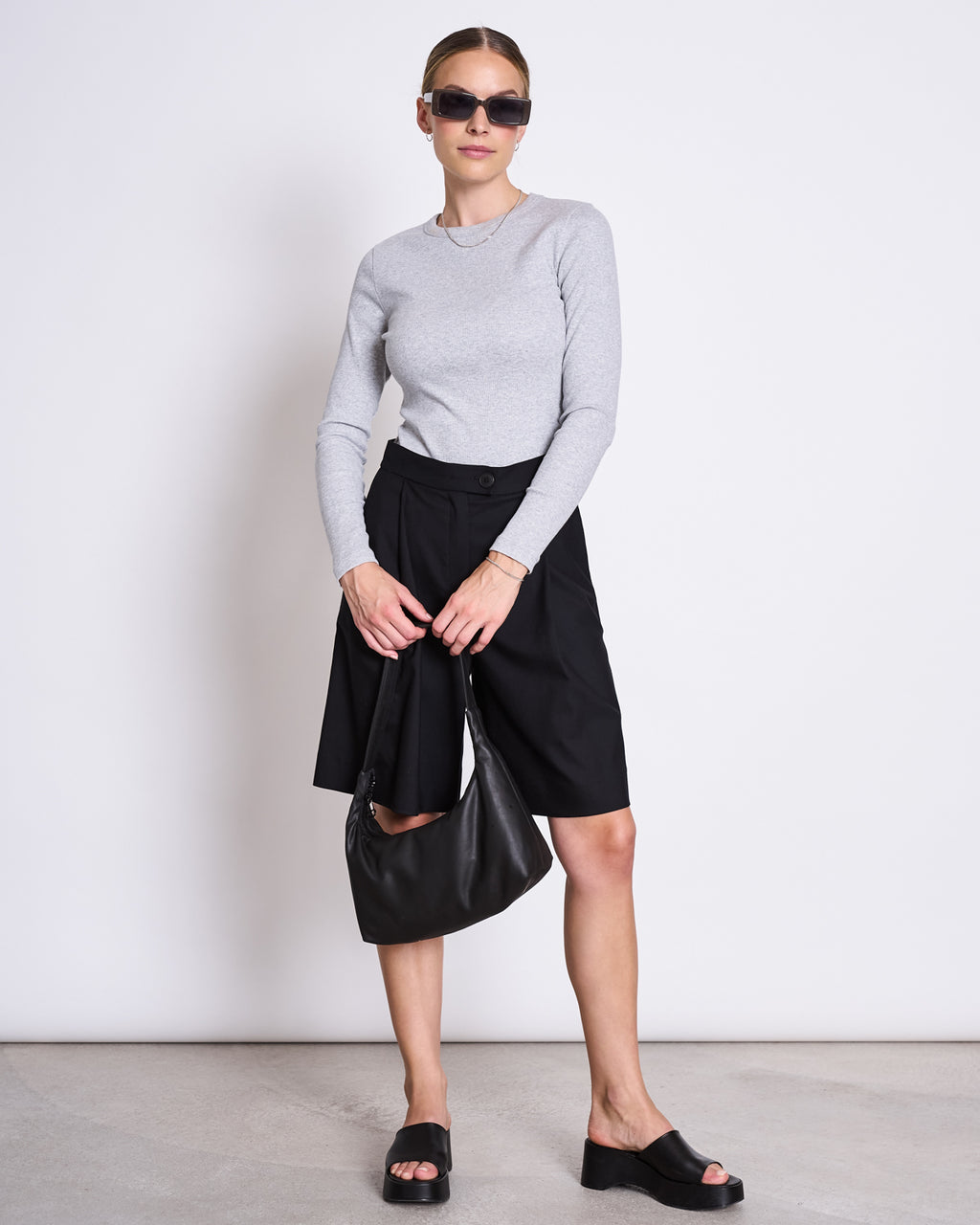 A woman wears the jan-n-june RIB LONGSLEEVE MURI LIGHT GREY GOTS top with black shorts, platform sandals, a black handbag, and black jacket—an effortlessly layered look against a white wall.