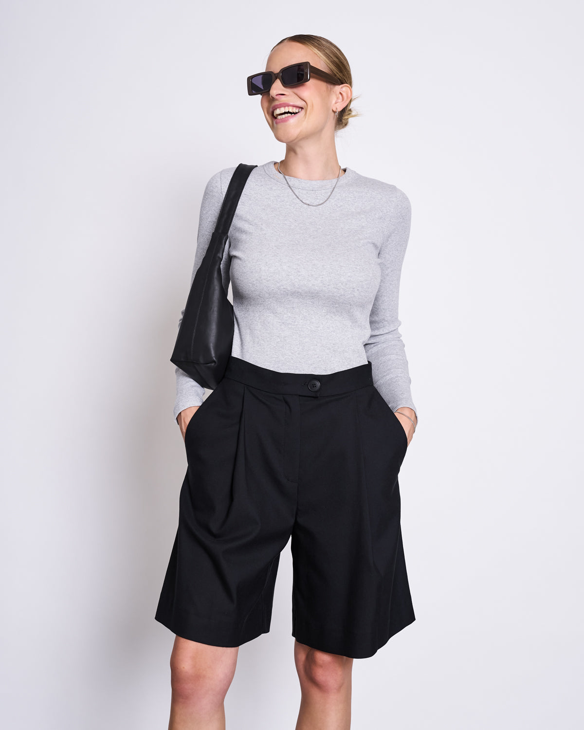 A smiling woman models the jan-n-june RIB LONGSLEEVE MURI LIGHT GREY GOTS top, styled with black knee-length shorts, dark sunglasses, a necklace, and a black shoulder bag against a white background with her hands in her pockets.