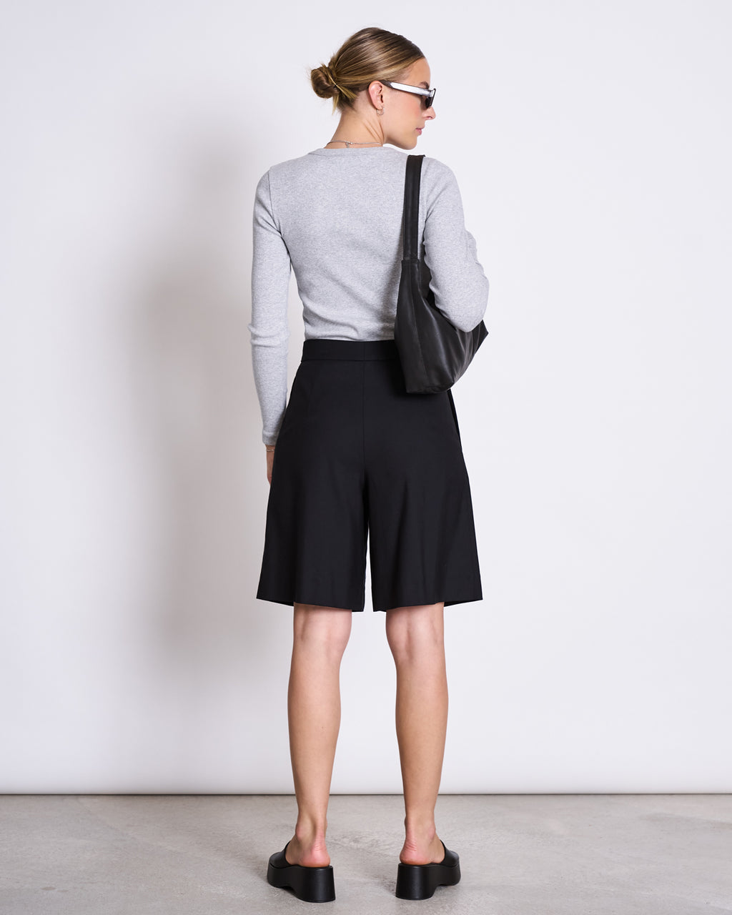 A woman faces away from the camera, wearing jan-n-june’s RIB LONGSLEEVE MURI LIGHT GREY GOTS—a slim, light gray layering top—paired with black knee-length shorts, platform sandals, sunglasses, and a black shoulder bag.