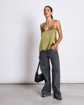 A woman with her dark hair pulled back wears the jan-n-june SLIP TOP MAUD WOODY OLIVE, styled with loose gray jeans, black sandals, a black bag, and minimal jewelry against a plain background.