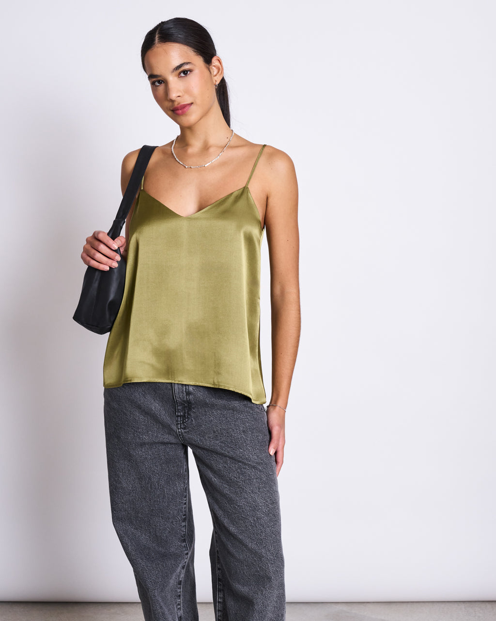 A woman models the jan-n-june SLIP TOP MAUD WOODY OLIVE, a shiny satin camisole with a wide V-neckline. She pairs it with loose gray jeans and a black shoulder bag, standing against a white background with her dark hair pulled back.