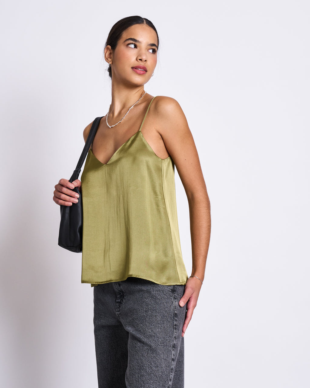 A woman in the SLIP TOP MAUD WOODY OLIVE by jan-n-june, featuring a shiny wide V-neck and crafted from sustainable ENKA® Viscose, pairs it with gray jeans and a black bag while standing against a white background, looking to the side.