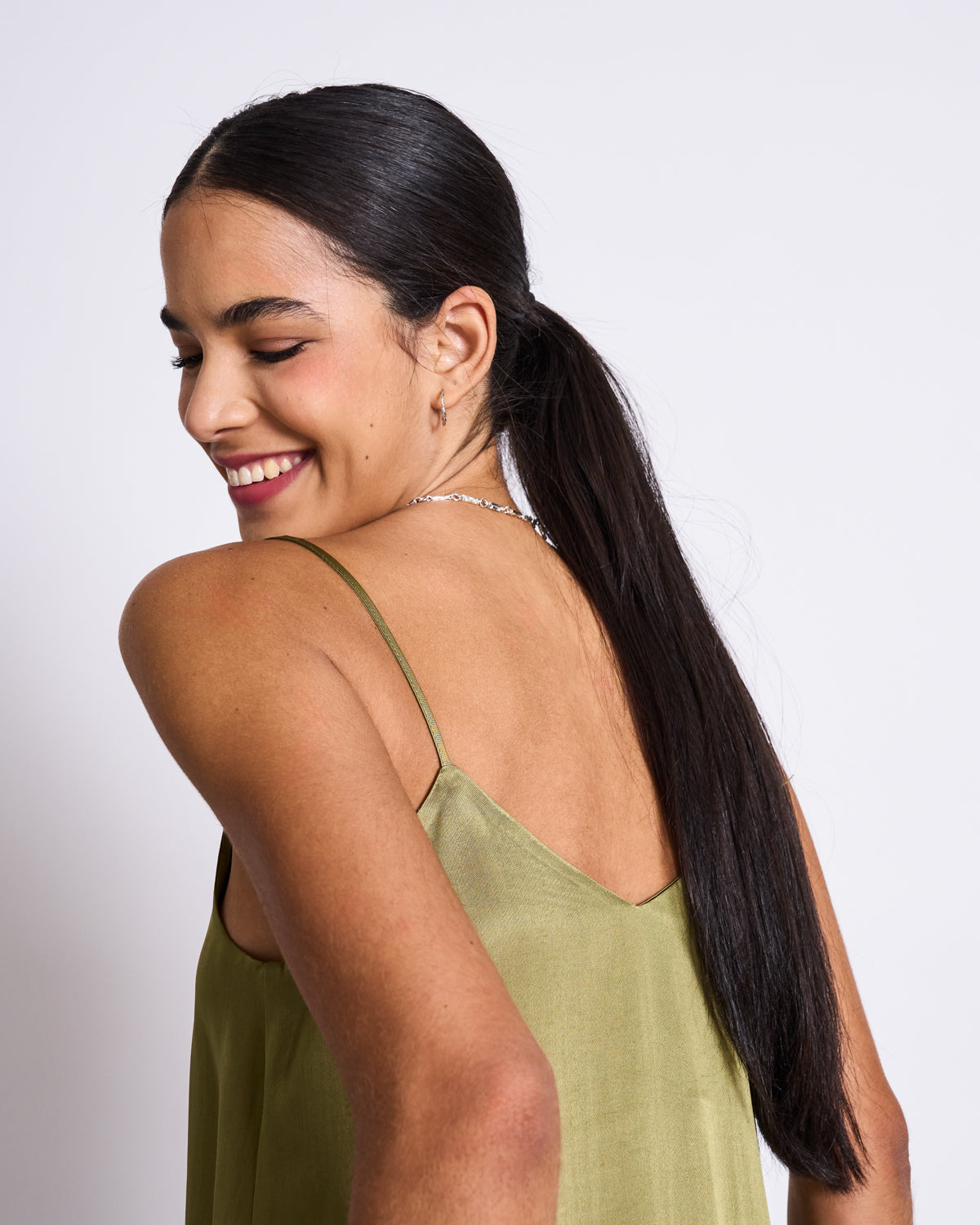 A woman with long dark hair in a low ponytail smiles with her eyes closed, wearing the jan-n-june SLIP TOP MAUD WOODY OLIVE—a sleeveless top with a wide V-neckline—paired with a silver necklace, posed against a plain white background.