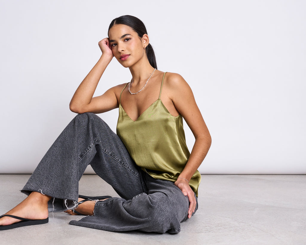 A woman with long dark hair sits on the floor against a plain light background, wearing the jan-n-june SLIP TOP MAUD WOODY OLIVE, paired with gray wide-leg jeans and black sandals, her chin resting on her hand.