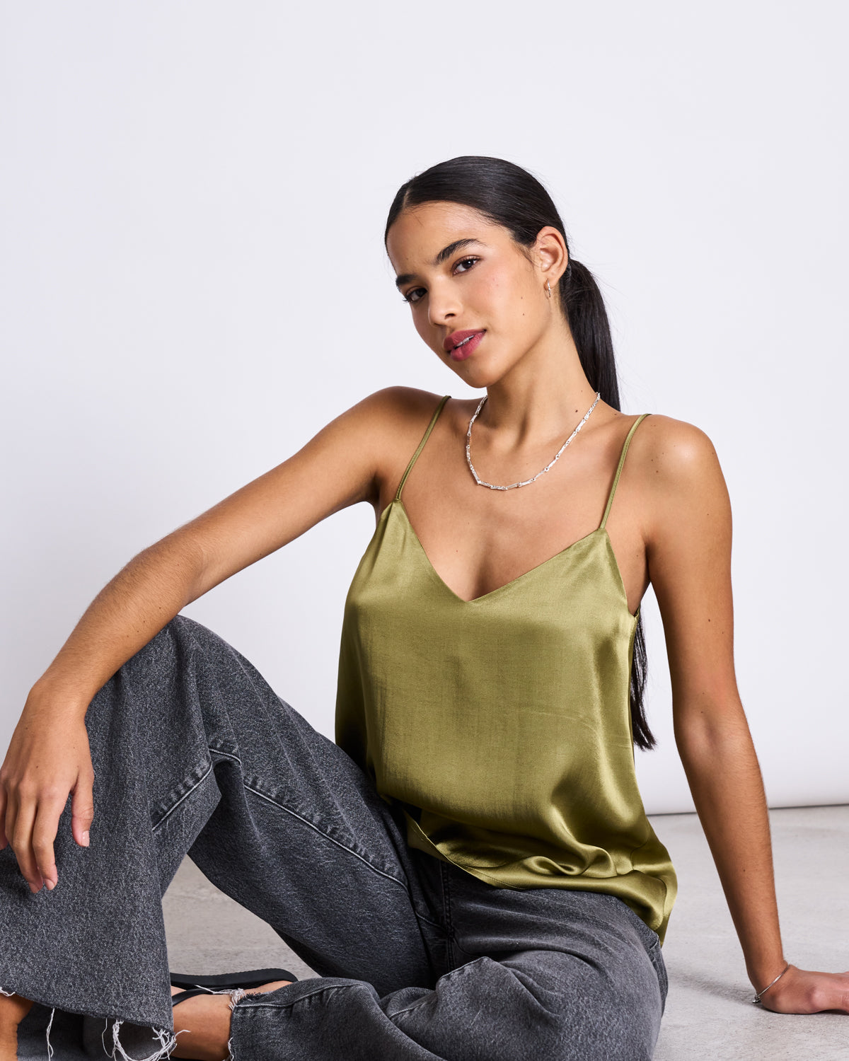 A woman with long dark hair in a low ponytail sits on the floor against a plain white background, wearing the jan-n-june SLIP TOP MAUD WOODY OLIVE and loose gray jeans, looking calmly at the camera.