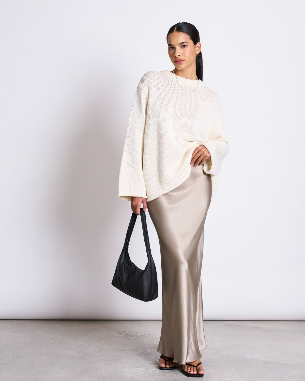 A woman stands against a plain background wearing the jan-n-june JUMPER SONA OFFWHITE GOTS, an oversized organic cotton knit sweater, paired with a long beige skirt. She holds a black bag, wears black sandals, and gazes confidently at the camera.