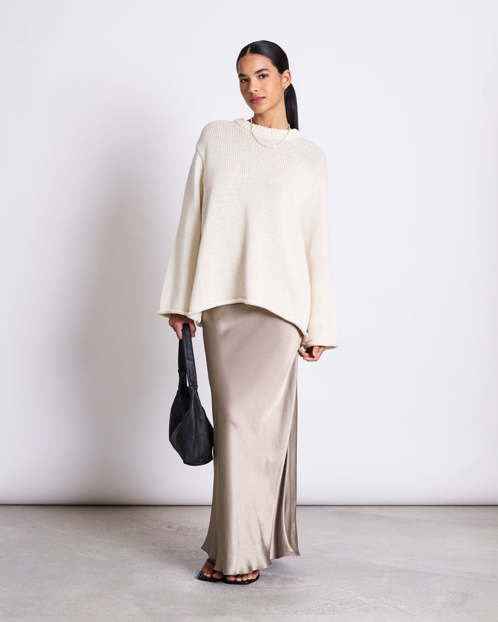 A woman with dark hair pulled back stands against a plain background, wearing the jan-n-june JUMPER SONA OFFWHITE GOTS—a 100% organic cotton oversized sweater—over a beige skirt, paired with sandals and a black handbag.