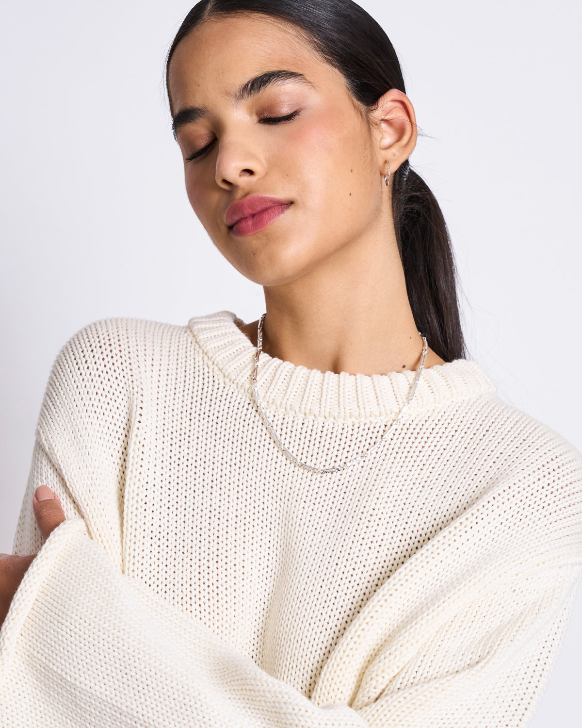 A woman with long dark hair in a low ponytail poses with eyes closed and arms crossed against a plain background, wearing the jan-n-june JUMPER SONA OFFWHITE GOTS, an oversized knit sweater made from 100% organic cotton.