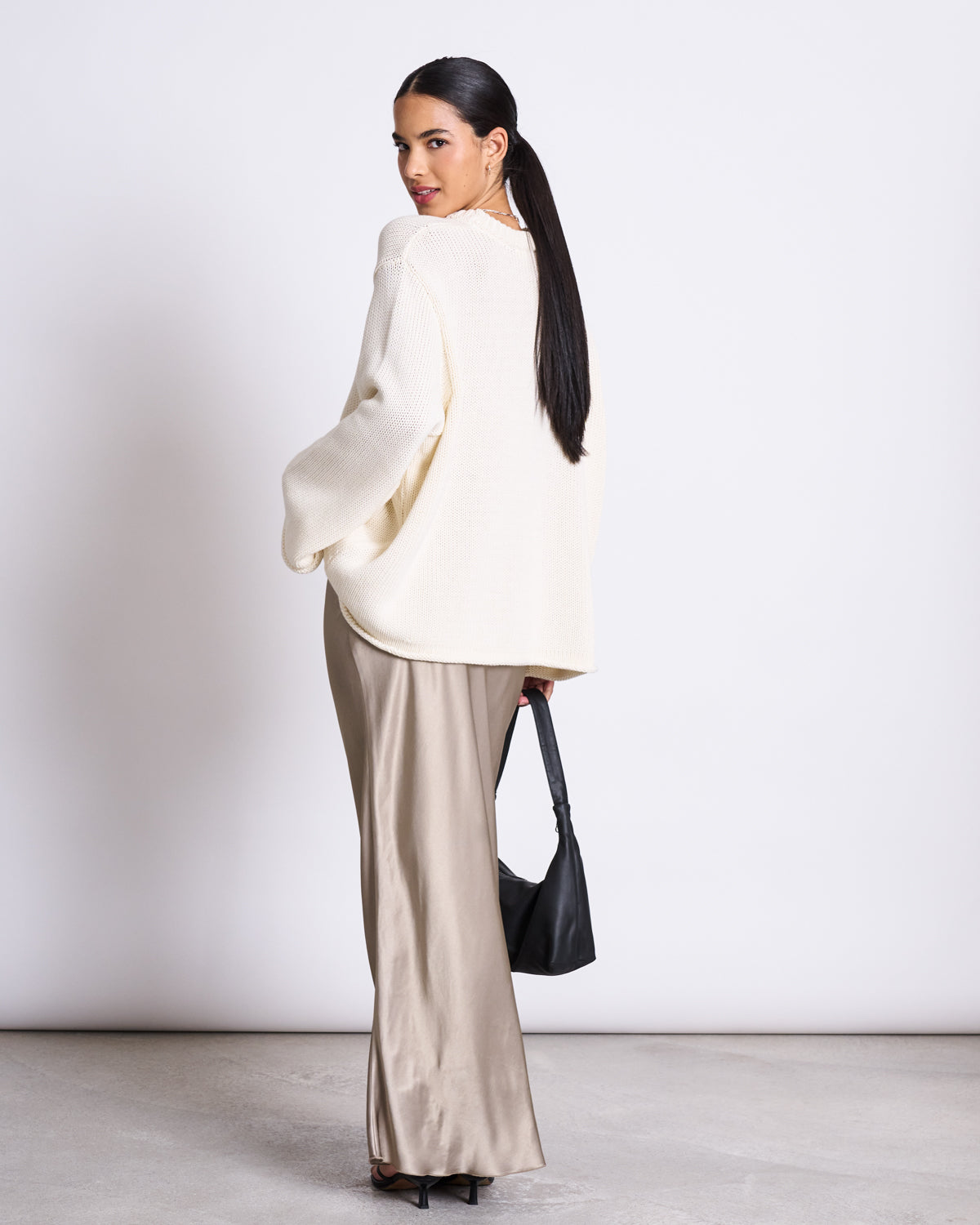 A woman with long dark hair poses against a white background, wearing the jan-n-june JUMPER SONA OFFWHITE GOTS—an oversized cream knit sweater made from 100% organic cotton—with beige satin pants and a black handbag.