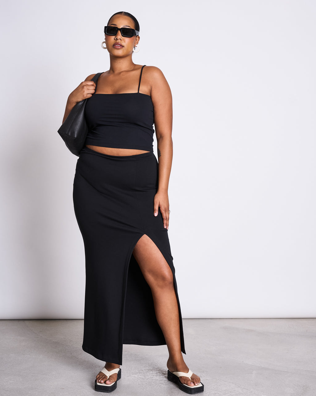 A woman stands confidently against a plain light background, wearing sunglasses, a black sleeveless crop top, sandals, a black shoulder bag, and the jan-n-june MAXI SKIRT LUSTA BLACK with a thigh-high slit.