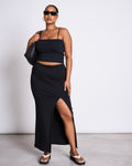 A woman stands confidently against a plain light background, wearing sunglasses, a black sleeveless crop top, sandals, a black shoulder bag, and the jan-n-june MAXI SKIRT LUSTA BLACK with a thigh-high slit.