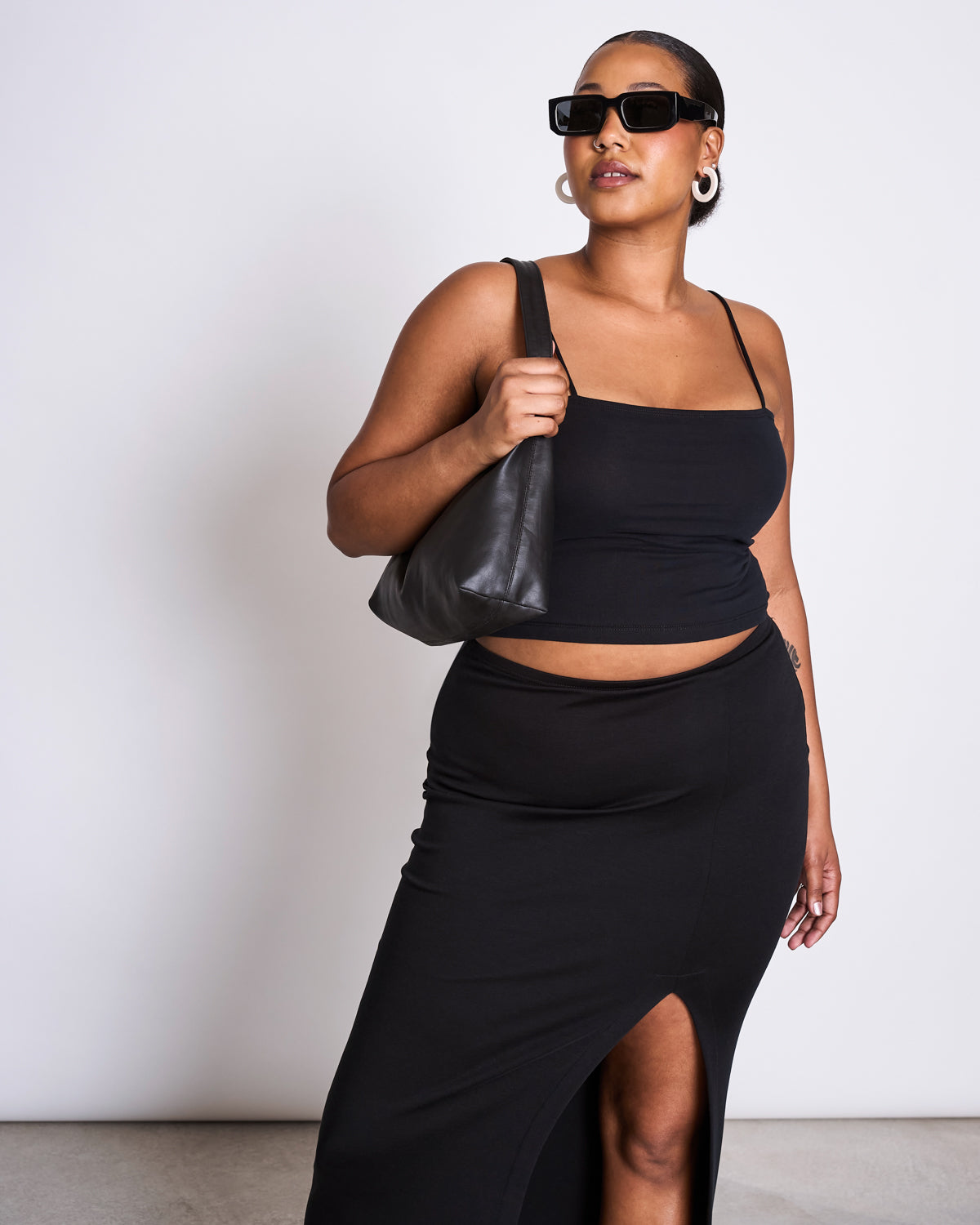 A woman wears the jan-n-june MAXI SKIRT LUSTA BLACK with a sleeveless top, sunglasses, hoop earrings, and a black shoulder bag, posing against a plain white background.