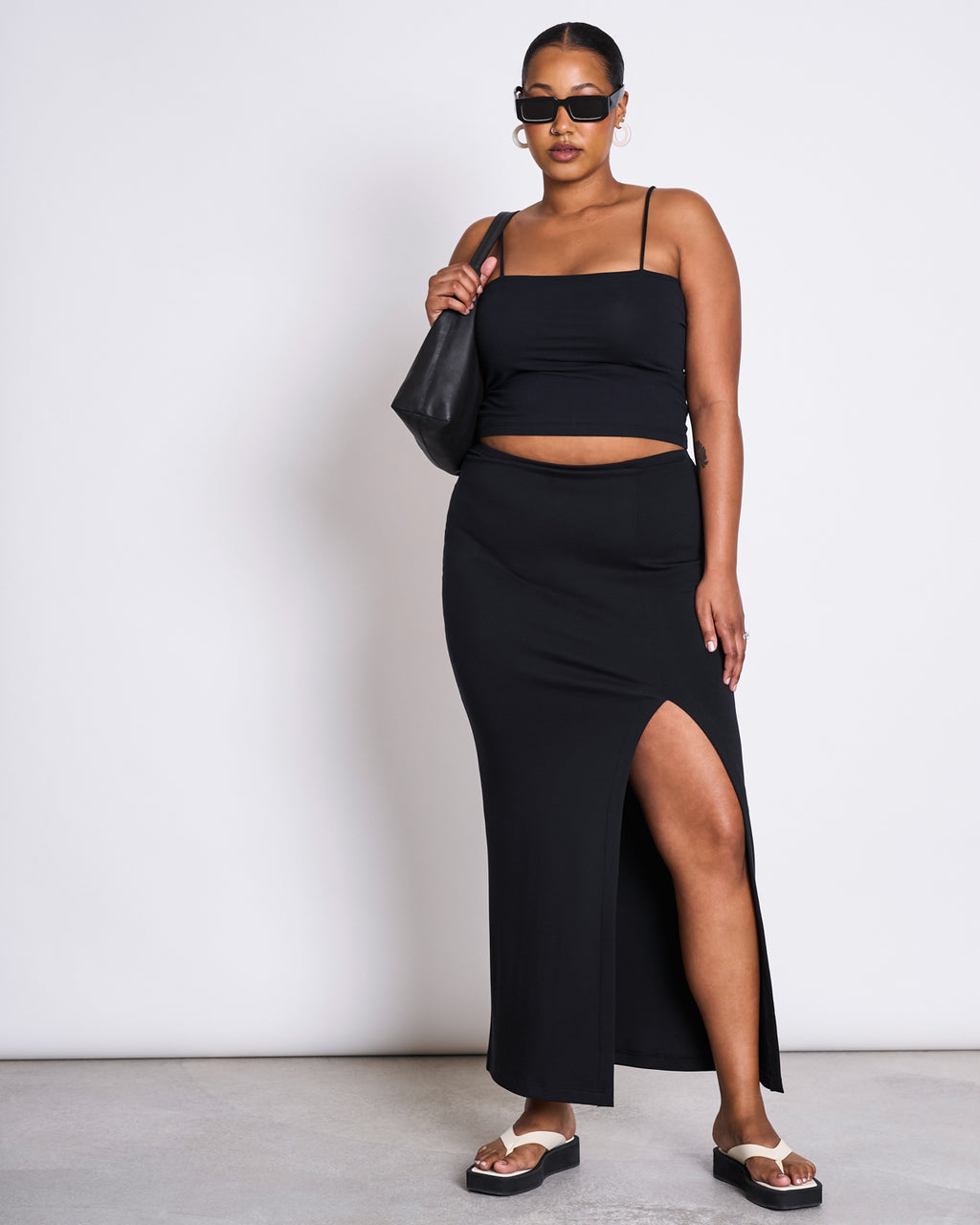 A woman stands against a white background wearing the jan-n-june MAXI SKIRT LUSTA BLACK, a black crop top, sunglasses, hoop earrings, black platform sandals, and carrying a black shoulder bag.