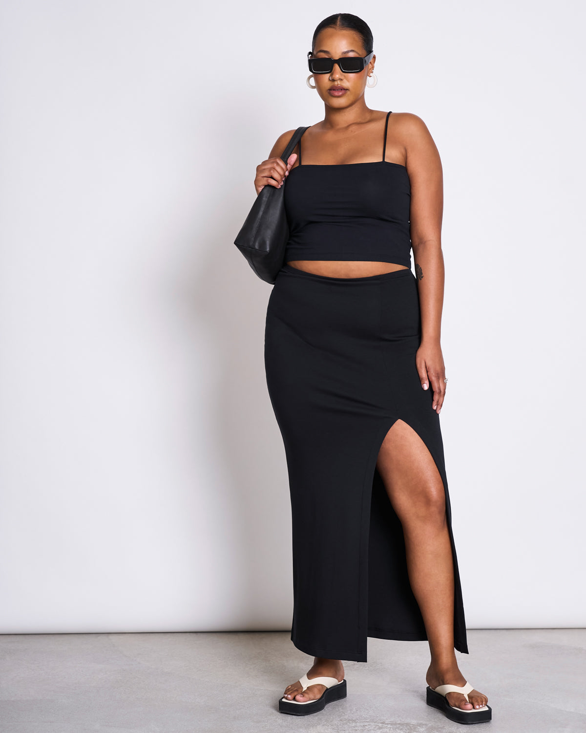 A woman stands against a white background wearing the jan-n-june MAXI SKIRT LUSTA BLACK, a black crop top, sunglasses, hoop earrings, black platform sandals, and carrying a black shoulder bag.
