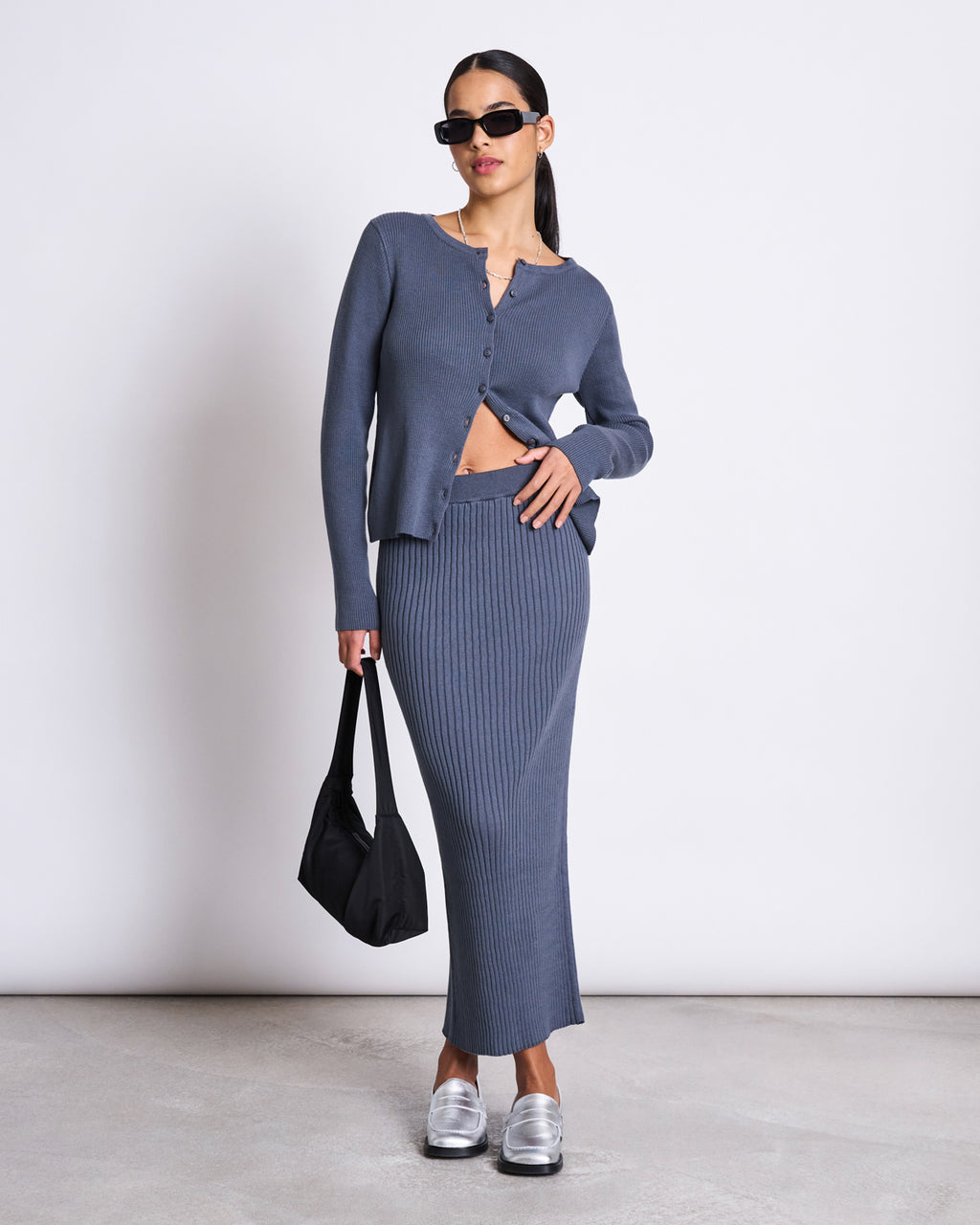 A woman wearing black sunglasses, a blue buttoned cardigan, and the jan-n-june MIDI KNIT SKIRT EBBA TURBULENCE GOTS poses with one hand on her hip and holds a black bag indoors on a light floor, paired with metallic shoes.