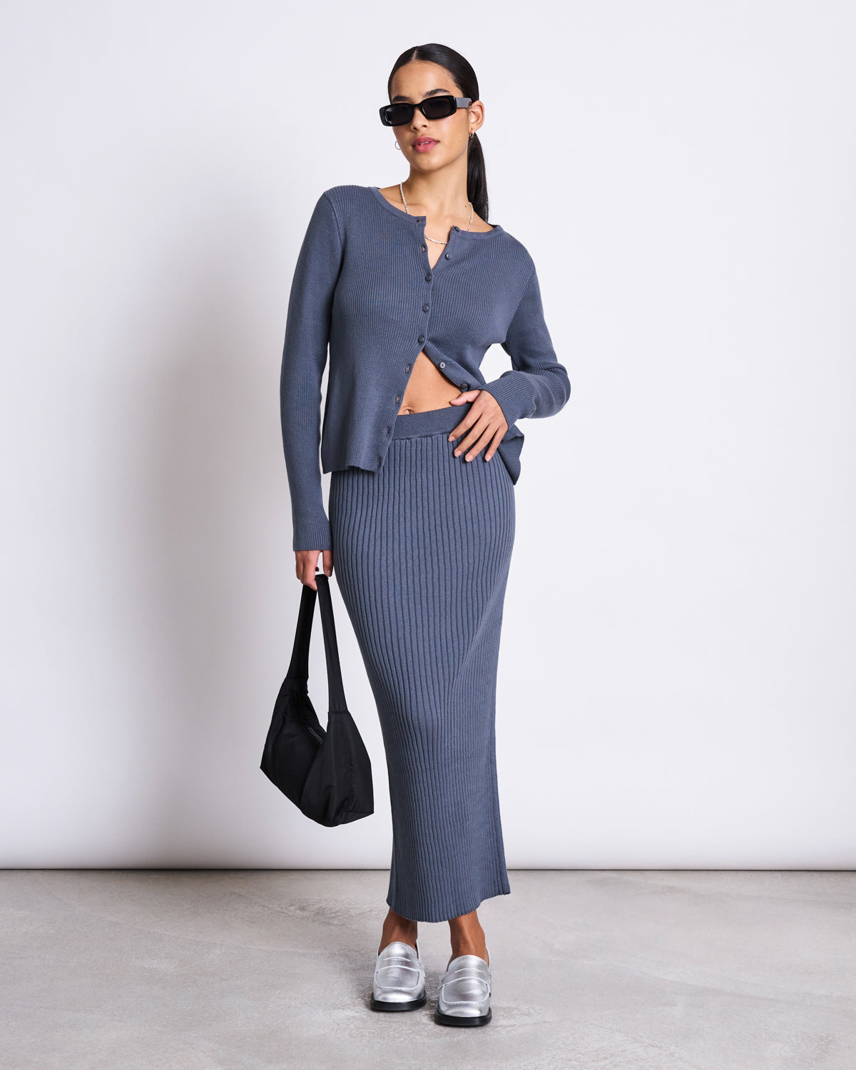 A woman wearing black sunglasses, a blue buttoned cardigan, and the jan-n-june MIDI KNIT SKIRT EBBA TURBULENCE GOTS poses with one hand on her hip and holds a black bag indoors on a light floor, paired with metallic shoes.