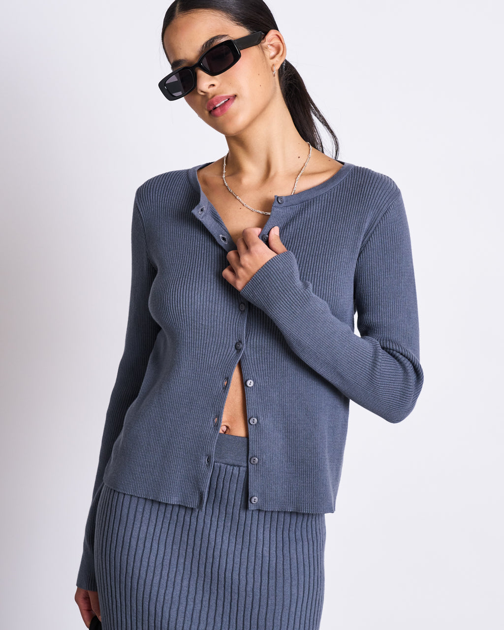 A woman wearing the jan-n-june CREWNECK CARDIGAN GLIMT TURBULENCE GOTS in blue-gray and a matching skirt poses against a white background, her dark hair pulled back, accessorized with black sunglasses and a delicate necklace.