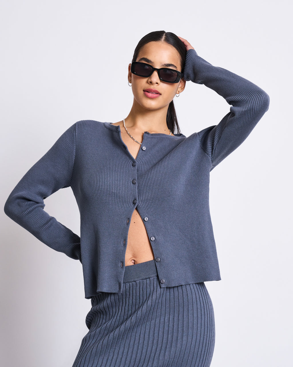 A woman poses with one hand on her hip in the jan-n-june MIDI KNIT SKIRT EBBA TURBULENCE GOTS, paired with a matching ribbed dark gray cardigan. She accessorizes with black sunglasses and small hoop earrings.