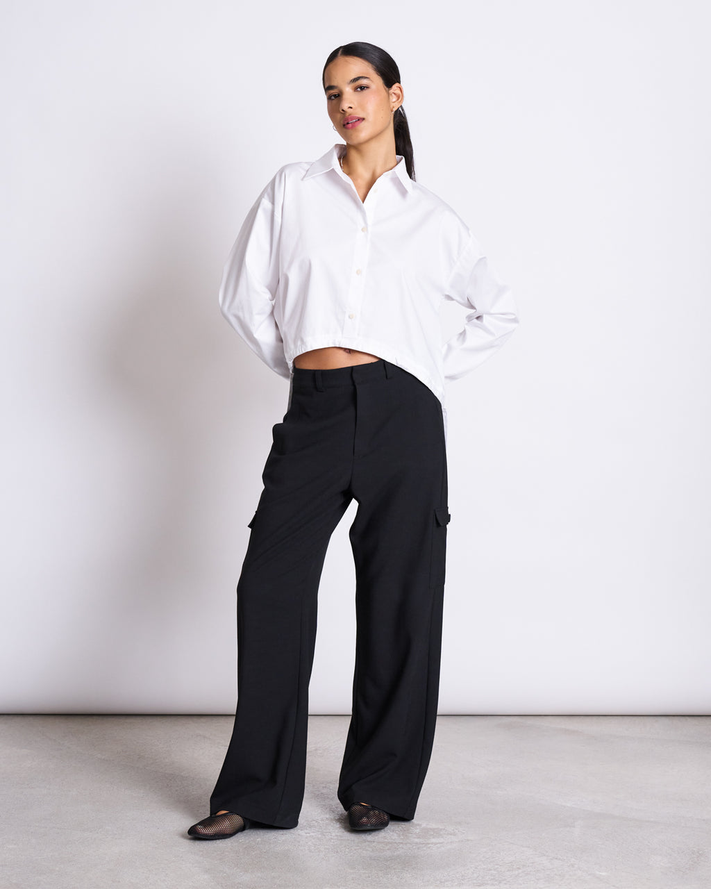 A woman stands against a plain background wearing the jan-n-june DRAWSTRING SHIRT ISTED WHITE, an oversized fit with dropped shoulders, paired with loose black pants and black flats. Her dark hair is pulled back, exuding relaxed confidence.