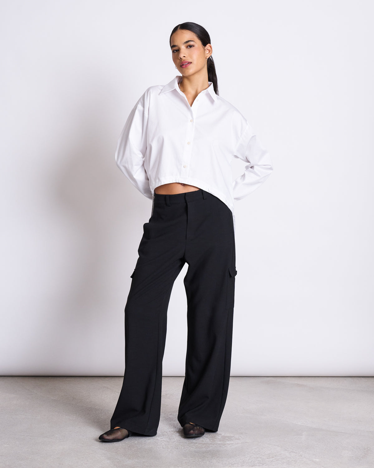 A woman stands against a plain background wearing the jan-n-june DRAWSTRING SHIRT ISTED WHITE, an oversized fit with dropped shoulders, paired with loose black pants and black flats. Her dark hair is pulled back, exuding relaxed confidence.