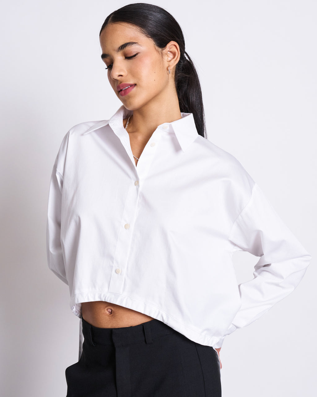 A woman with long dark hair in a low ponytail wears the jan-n-june DRAWSTRING SHIRT ISTED WHITE, a white oversized long-sleeved cropped button-up, paired with black pants. She stands with one hand on her hip, eyes closed and smiling slightly.