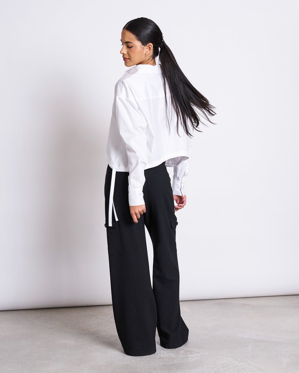 A woman with long dark hair wears the jan-n-june DRAWSTRING SHIRT ISTED WHITE and black wide-leg pants, standing on a light gray floor facing away and looking over her shoulder against a plain white background.