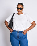 A woman wearing jan-n-june’s BRUSHED T-SHIRT BOY PLAIN WHITE GOTS, oversized sunglasses, and blue jeans stands against a plain background with a black shoulder bag and her hands on her hips.