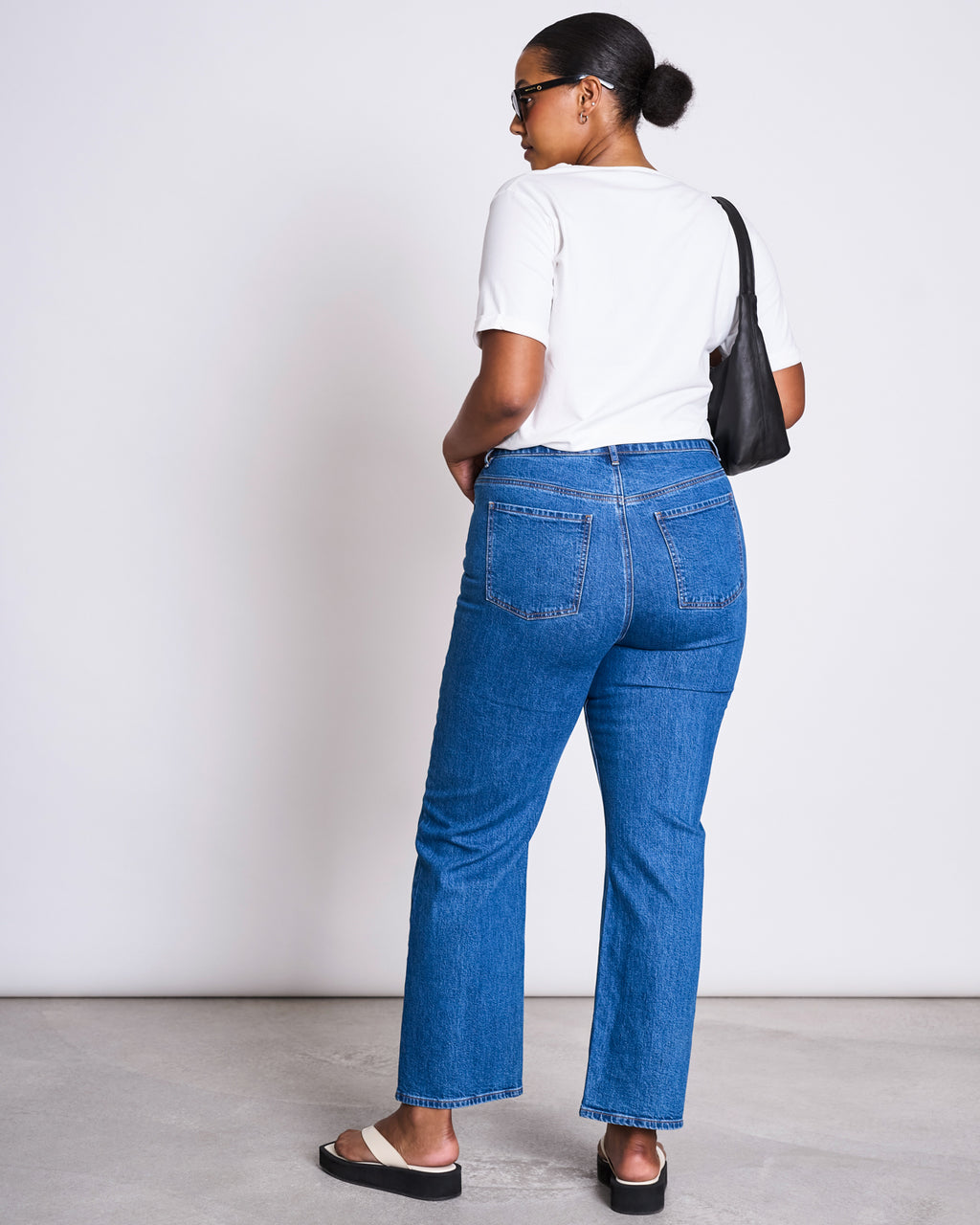 A woman with her hair in a bun faces away, wearing the jan-n-june BRUSHED T-SHIRT BOY PLAIN WHITE GOTS, blue jeans, black sandals, sunglasses, and carrying a black shoulder bag against a plain light background.