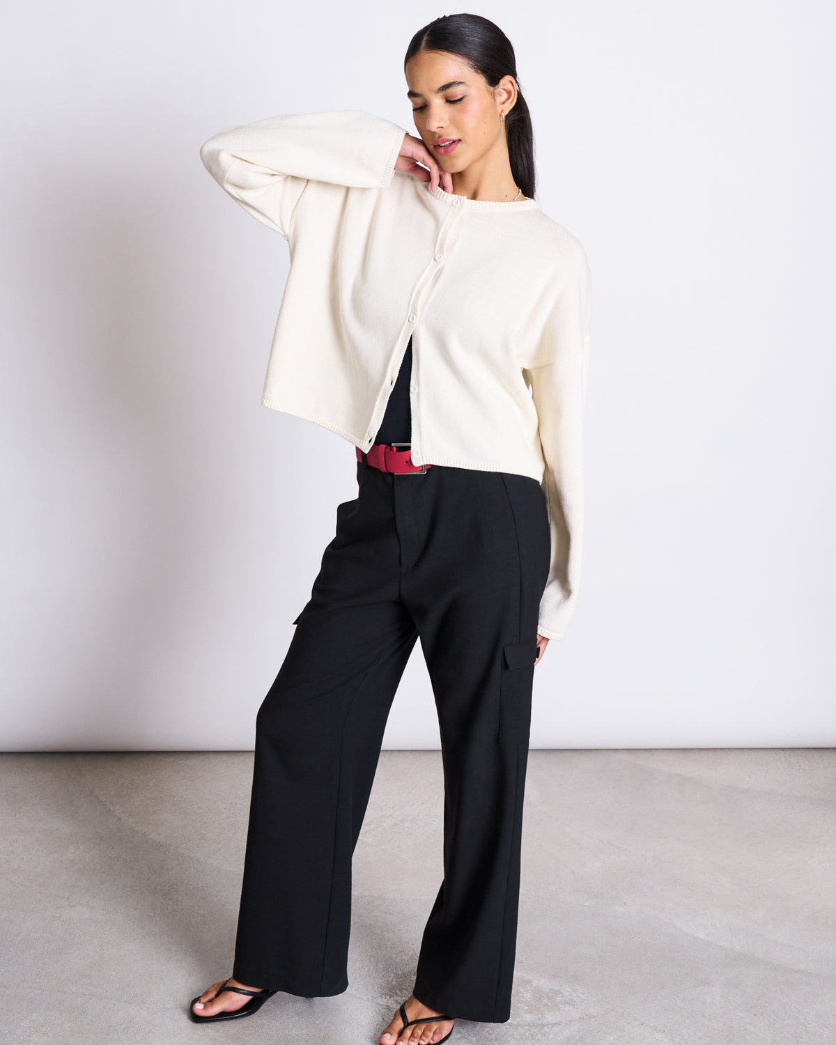 A woman stands in a relaxed pose wearing the jan-n-june LIGHT CARDIGAN ASAS OFFWHITE GOTS over a black top, wide-leg black pants with a red belt, and black sandals, against a plain light background.