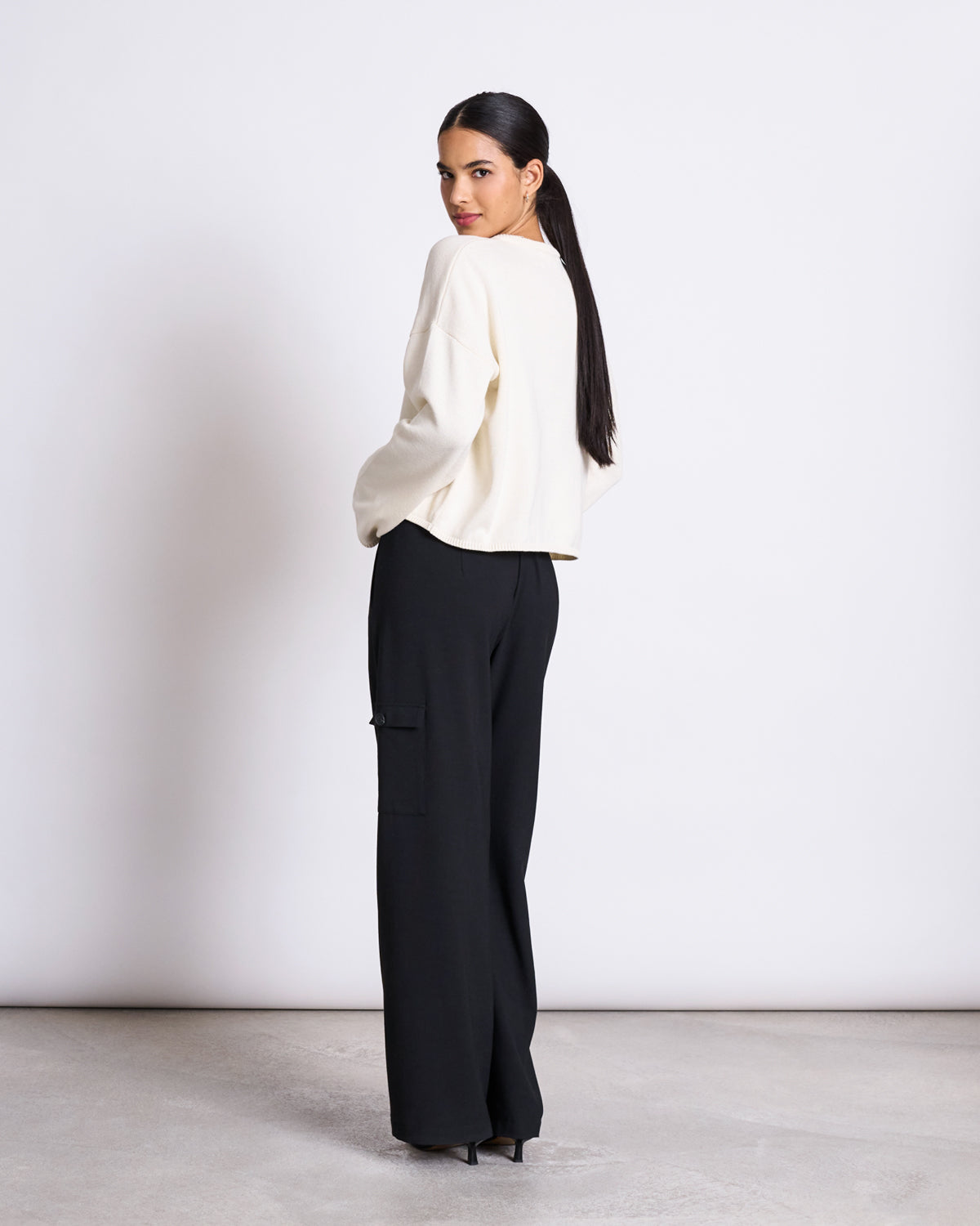 A woman with long dark hair stands sideways, looking over her shoulder. She wears the jan-n-june LIGHT CARDIGAN ASAS OFFWHITE GOTS with black wide-leg trousers and black heels against a plain white background.