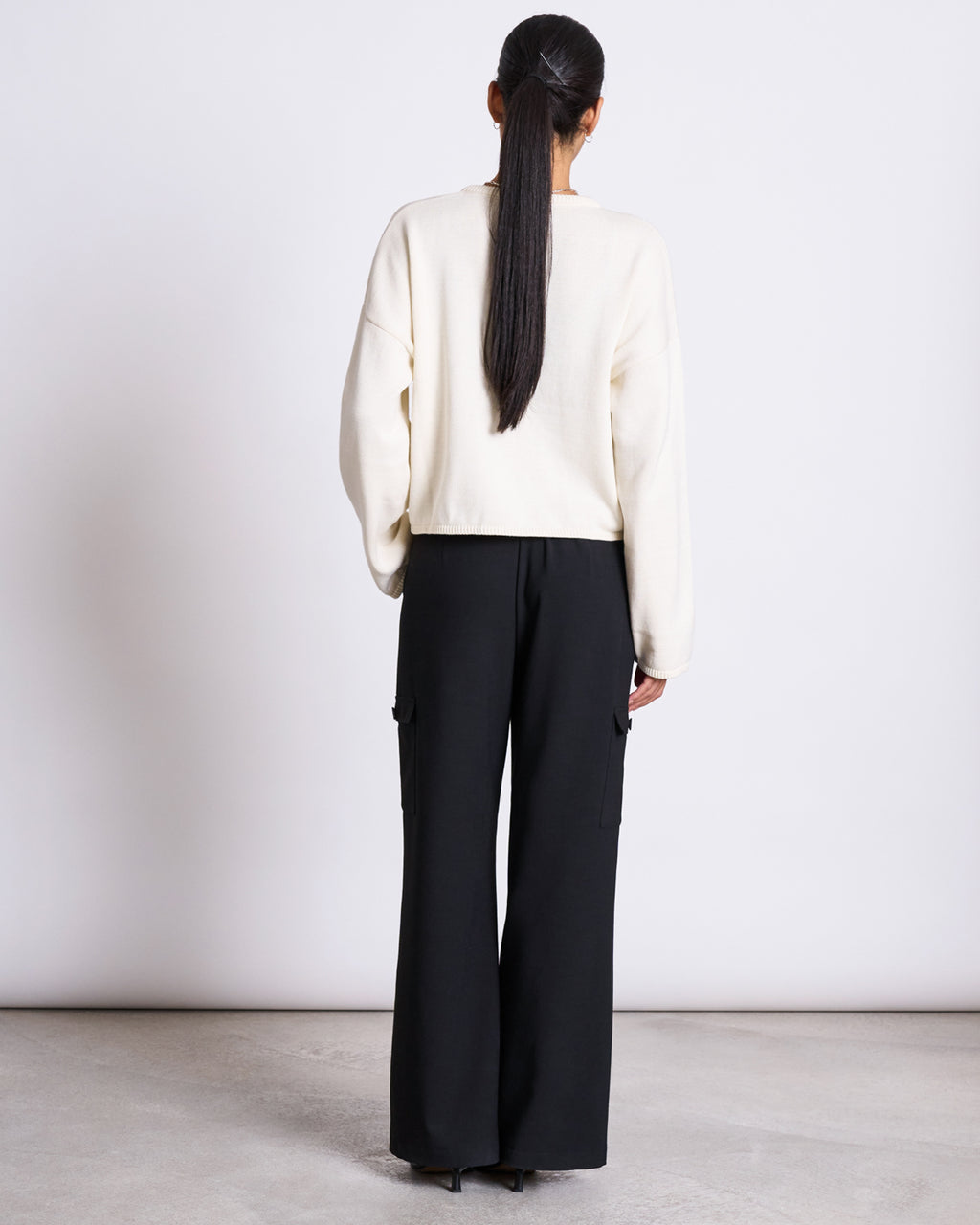 A person with long dark hair tied back is seen from behind, wearing the jan-n-june LIGHT CARDIGAN ASAS OFFWHITE GOTS and loose black cargo trousers, standing against a plain white background.