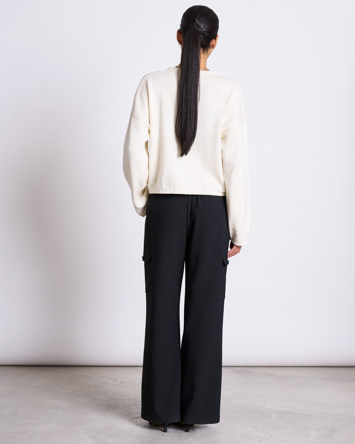 A person with long dark hair tied back is seen from behind, wearing the jan-n-june LIGHT CARDIGAN ASAS OFFWHITE GOTS and loose black cargo trousers, standing against a plain white background.