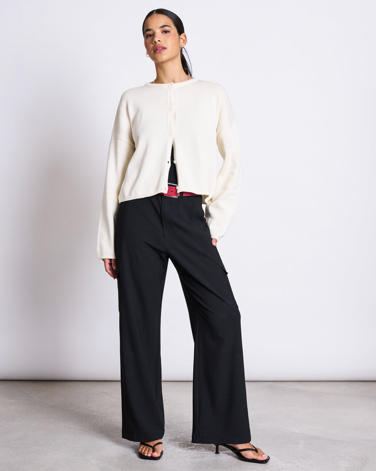 A woman poses confidently with one hand in her pocket, wearing the jan-n-june LIGHT CARDIGAN ASAS OFFWHITE GOTS over a black top, paired with black wide-leg pants, a red belt, and black heeled sandals against a plain background.