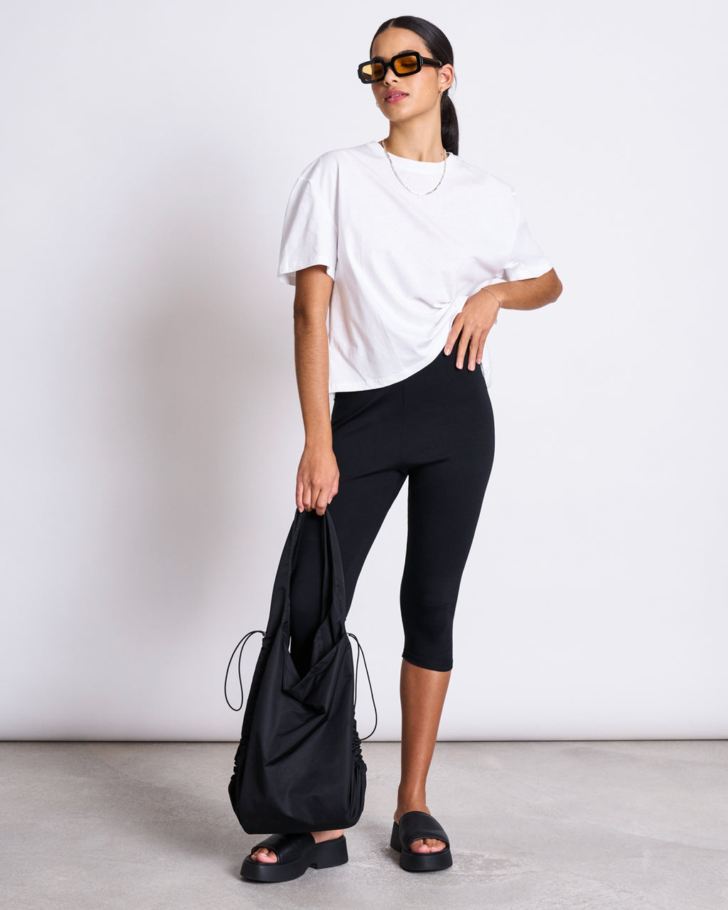 A woman stands against a plain background wearing the jan-n-june BOXY T-SHIRT TEMEL WHITE, black knee-length leggings, black platform sandals, sunglasses, and holding a black tote bag.
