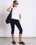 A woman in sunglasses wears the oversized jan-n-june BOXY T-SHIRT TEMEL WHITE, made from organic cotton, paired with black cropped leggings and platform slides. She stands against a plain background holding a black shoulder bag.
