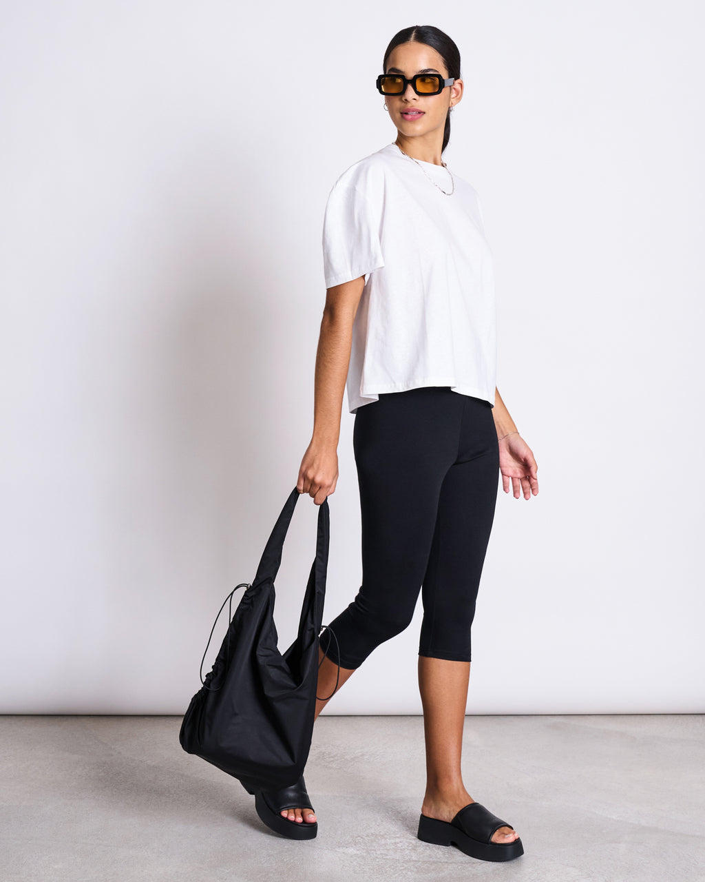 A woman in sunglasses wears the jan-n-june BOXY T-SHIRT TEMEL WHITE, black capri leggings, and black slide sandals, carrying a large black bag. She stands on a concrete floor against a plain white background.